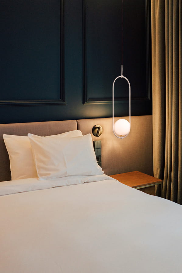 Lampa wisząca Malta 1xE14 w kolorze białym, umieszczona nad łóżkiem. Posiada prostą, okrągłą kulę świetlną w metalowej ramie, zawieszona na cienkim przewodzie. W tle widoczna ciemna ściana i zasłony, co tworzy elegancką aranżację sypialni.