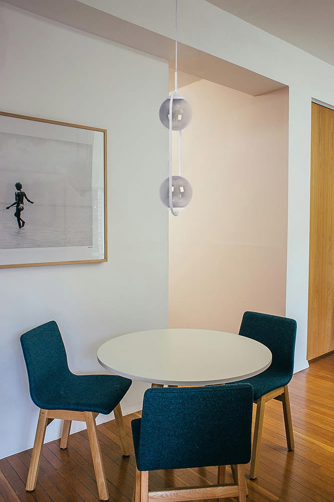 Lampa wisząca Arlena 2xE14 w kolorze białym i grafitowym, z dwoma szklanymi kulami, zawieszona nad okrągłym stołem w jasnym wnętrzu. Otoczenie to drewniana podłoga i niebieskie krzesła.