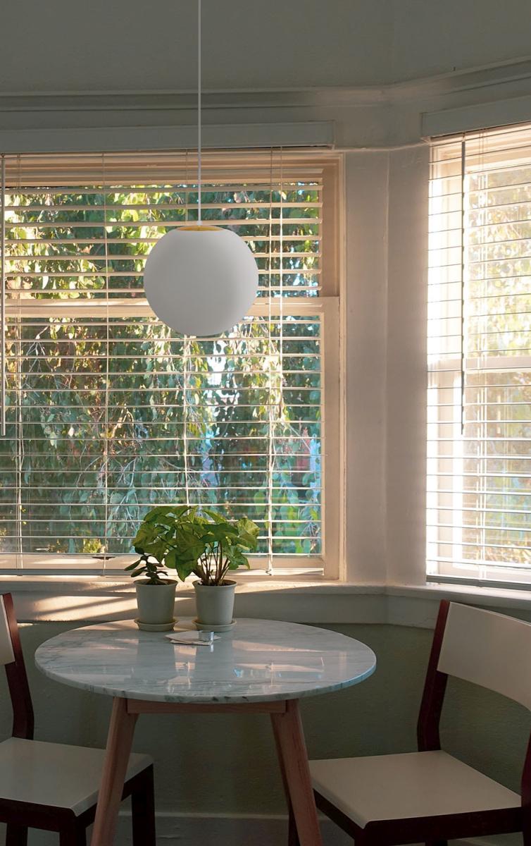 Biała lampa wisząca Elmo D30 z metalowym wykończeniem w kolorze złotym, o okrągłym kształcie, zawieszona na długim kablu. Umieszczona nad okrągłym, marmurowym stołem w jasnym wnętrzu z roślinami doniczkowymi.