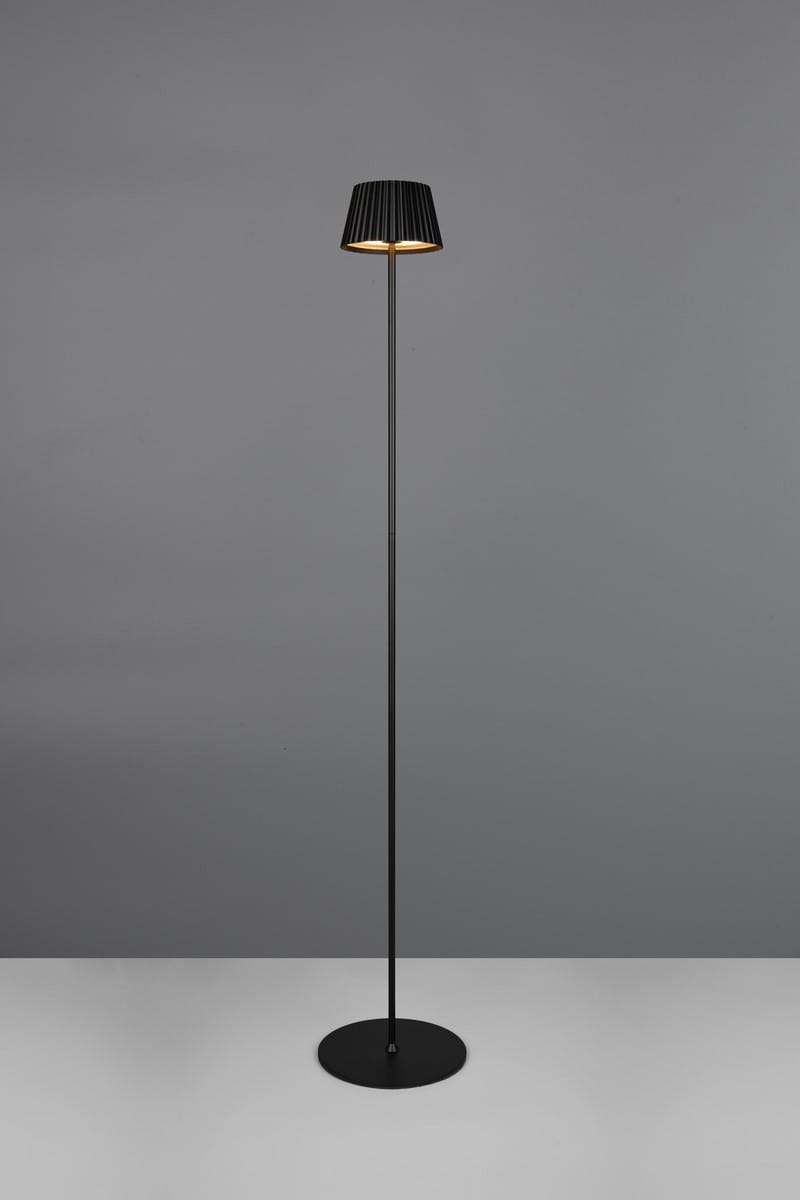 Lampa stojąca zewnętrzna Suarez w kolorze czarnym, z metalową podstawą i kloszem w formie plisowanej. Ujęcie frontalne, lampa ustawiona w pozycji stojącej, z widocznym źródłem światła LED o mocy 1,5W. Tło w odcieniach szarości.