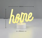 Lampa stołowa Casa LED o białym kolorze z napisem home w formie żółtego neonu. Produkt ma wymiary 240 mm szerokości i 165 mm wysokości, osadzony na okrągłej podstawie o średnicy 85 mm. Tło w odcieniu szarym.