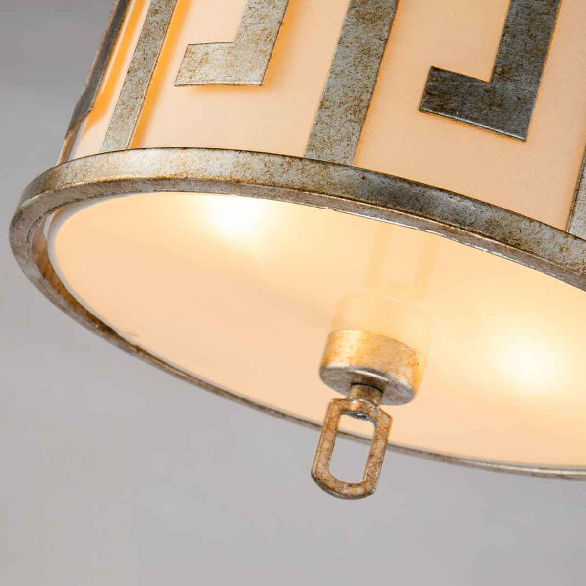 Lampa wisząca Lemuria w kolorze starego złota, z okrągłym abażurem osłoniętym jasnym materiałem. Widoczny detal mocowania oraz metalowe wykończenie. Produkt przeznaczony na dwie żarówki E27. Umiejscowiona w górnej części zdjęcia.