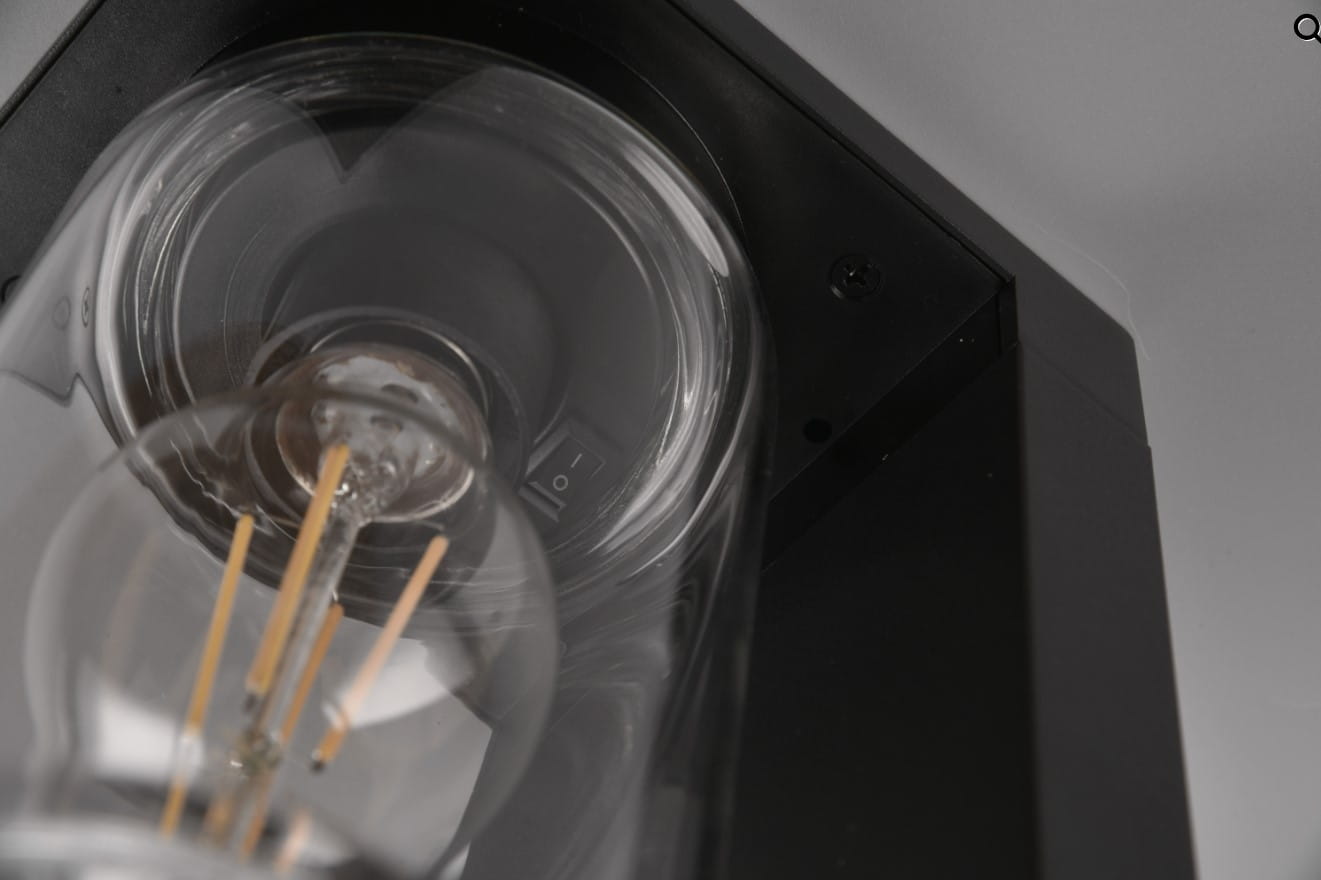 Czarna lampa zewnętrzna Ardila H100 LED z czujnikiem zmierzchu, stojąca, widok z góry. Wewnątrz przezroczysta żarówka o widocznych elementach, włącznik na obudowie. Prosta, nowoczesna konstrukcja, idealna do oświetlenia przestrzeni na zewnątrz.