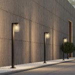 Czarna lampa zewnętrzna Ardila H100 z czujnikiem zmierzchu, stojąca, z przezroczystym kloszem. Ujęcie z boku, pokazujące trzy lampy ustawione wzdłuż ściany, na tle gładkiej, betonowej powierzchni, z białym żwirem na ziemi.