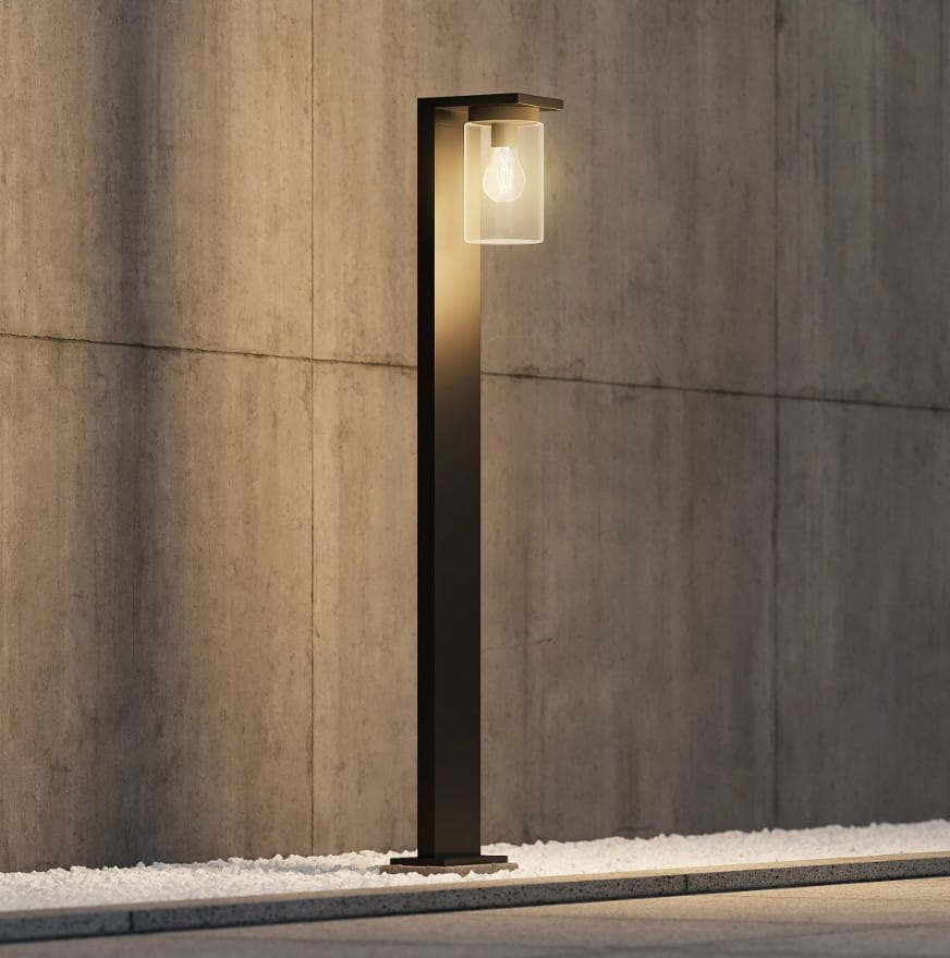 Stojąca lampa zewnętrzna Ardila H100 w kolorze czarnym, z przezroczystym kloszem i żarówką LED. Ujęcie z boku, na tle betonowej ściany. Wyposażona w czujnik zmierzchu, emituje ciepłe światło, umieszczona na płaskiej podstawie.