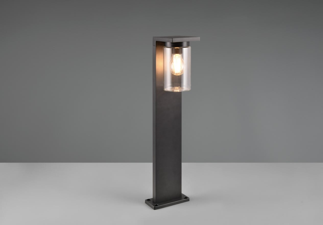 Czarna lampa zewnętrzna stojąca Ardila H65 z przezroczystym kloszem, wyposażona w czujnik zmierzchu. Ujęcie frontalne, lampa na prostokątnej podstawie, z widoczną żarówką wewnątrz klosza. Tło szare, minimalistyczne.