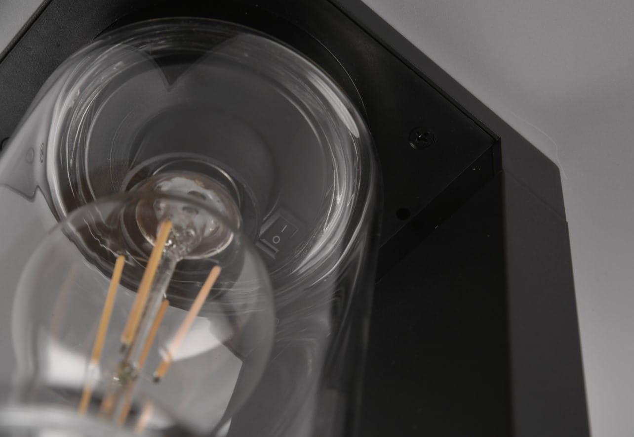 Czarna lampa zewnętrzna Ardila H65 LED z czujnikiem zmierzchu, stojąca. Widać szklany klosz z żarówką oraz przełącznik na obudowie. Produkt ukazany w zbliżeniu, eksponujący detale konstrukcji.