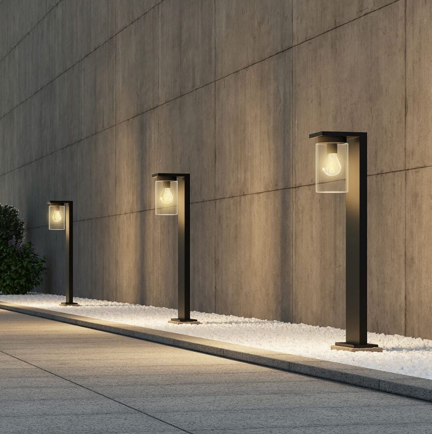 Czarna lampa zewnętrzna Ardila H65 z czujnikiem zmierzchu, stojąca, z przezroczystym kloszem, umieszczona przy ścianie w nowoczesnym otoczeniu. Widoczne trzy lampy na ścieżce, oświetlające otoczenie.