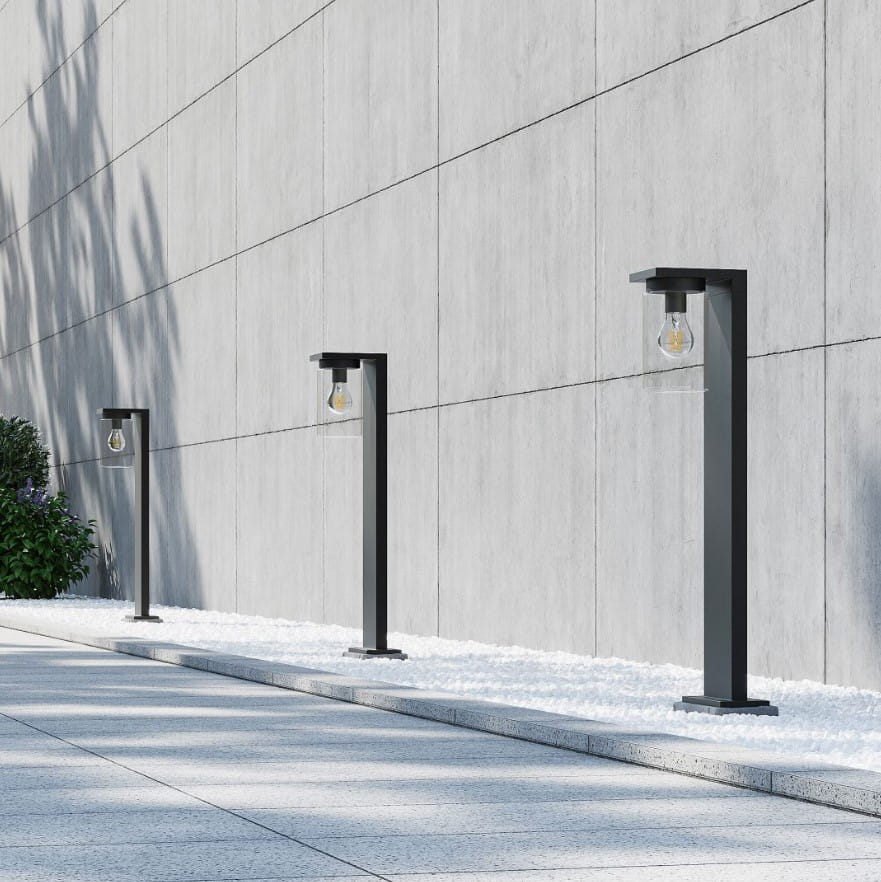 Czarna lampa zewnętrzna stojąca Ardila H65 z czujnikiem zmierzchu, z widoczną żarówką, umieszczona na prostokątnej podstawie. Ujęcie z boku, w otoczeniu kamiennej nawierzchni i zieleni.