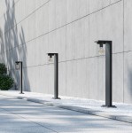 Czarna lampa zewnętrzna stojąca Ardila H65 z czujnikiem zmierzchu, z widoczną żarówką, umieszczona na prostokątnej podstawie. Ujęcie z boku, w otoczeniu kamiennej nawierzchni i zieleni.