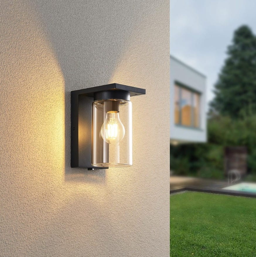 Kinkiet zewnętrzny Ardila 1-punktowy w kolorze czarnym z czujnikiem zmierzchu. Oświetlenie w formie szklanej osłony z widoczną żarówką, montowane na ścianie. W tle przyjemne otoczenie z zielonym trawnikiem i budynkiem.