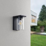 Kinkiet zewnętrzny Ardila, czarny, 1-punktowy z czujnikiem zmierzchu. Oświetlenie o prostokątnej podstawie z przezroczystym kloszem, umieszczony na jasnej ścianie, w tle trawnik i budynek. Klosz z żarówką widoczną w środku.