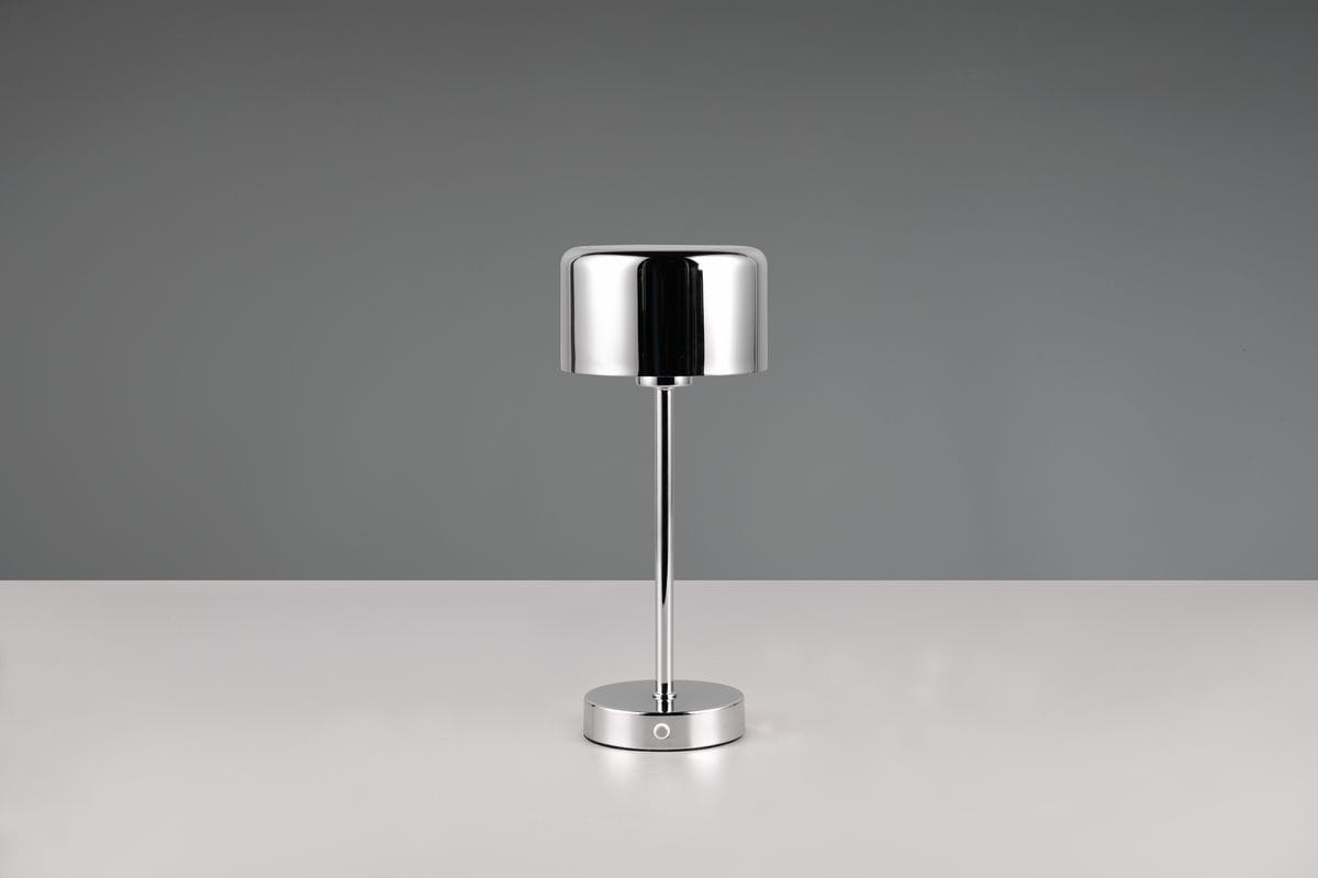Lampa stołowa Jeff w kolorze chromu, z cylindrycznym kloszem i prostą, metalową nogą. Ustawiona w pozycji stojącej na płaskiej powierzchni, z widocznym przyciskiem włącznika na podstawie. Idealna do nowoczesnych wnętrz.