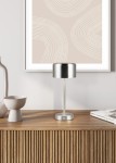 Lampa stołowa Jeff z metalowym stelażem w kolorze niklu, z cylindrycznym kloszem. Stoi na drewnianym stoliku obok ceramicznych akcesoriów. Tło jasne z abstrakcyjnym obrazem. Włącznik umieszczony na podstawie.