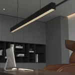 Czarna lampa wisząca Sigi II z diodą LED, o prostokątnym kształcie i matowej powierzchni, zawieszona nad biurkiem. W tle widoczne szare ściany oraz regały z książkami. Lampa emituje ciepłe światło, podkreślając nowoczesny styl wnętrza.