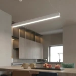 Lampa wisząca Sigi III w kolorze białym, z lampą LED 36W, zawieszona nad stołem w kuchni. Prosta, prostokątna forma z delikatnym podświetleniem, idealna do nowoczesnych wnętrz. W tle widoczne drewniane meble i naczynia.