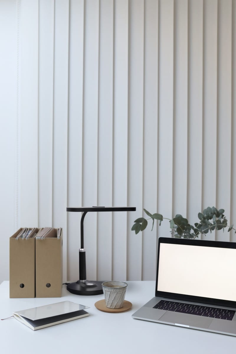 Czarna lampka biurkowa Dafi LED z 1 żarówką 10W, z minimalistycznym, okrągłym kloszem i smukłym ramieniem. Ustawiona na białym stole, obok laptopa i filiżanki na talerzyku. W tle teczki oraz rośliny doniczkowe.