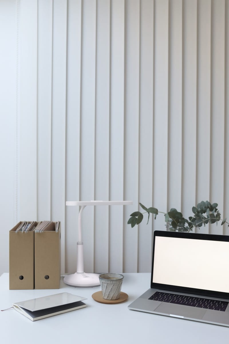 Biała lampka biurkowa Dafi LED z jedną żarówką 10W, o minimalistycznym designie. Ustawiona na biurku obok laptopa, filiżanki i teczek. Tło stanowi jasne, pionowe żaluzje, co podkreśla nowoczesny charakter aranżacji.