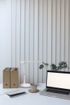 Biała lampka biurkowa Dafi LED z jedną żarówką 10W, o minimalistycznym designie. Ustawiona na biurku obok laptopa, filiżanki i teczek. Tło stanowi jasne, pionowe żaluzje, co podkreśla nowoczesny charakter aranżacji.