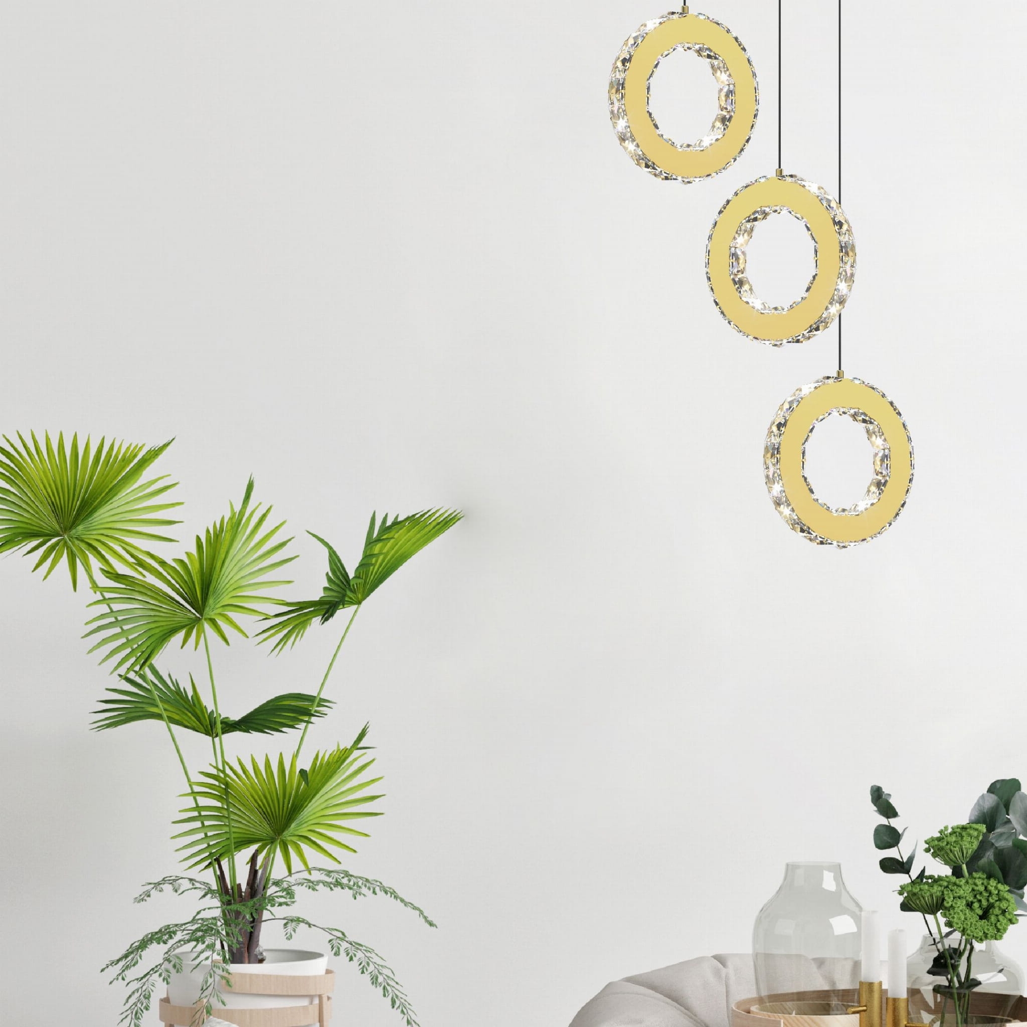 Lampa wisząca Girona 1xLED/10W w kolorze złotym, składająca się z trzech okrągłych elementów z przezroczystymi zdobieniami. Zawieszona na czarnych linkach w jasnym wnętrzu, obok rośliny doniczkowej.