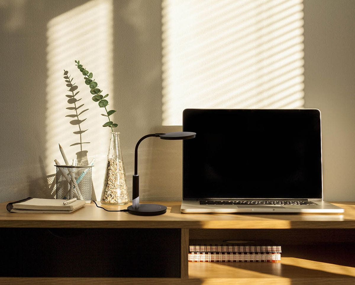 Lampka biurkowa Dino w kolorze czarnym z jedną diodą LED 10W, umieszczona na drewnianym biurku obok laptopa. W tle widoczne są rośliny w szklanych pojemnikach oraz notesy. Lampka ma smukły, nowoczesny kształt z okrągłą podstawą i elastycznym ramieniem.