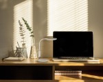 Biała lampka biurkowa Dino LED z jedną diodą 10W, umieszczona na drewnianym biurku obok laptopa. Lampka ma prostą, zakrzywioną podstawę i głowicę. W tle widoczne rośliny w wazonie oraz notes. Światło rzuca delikatne cienie na ścianę.