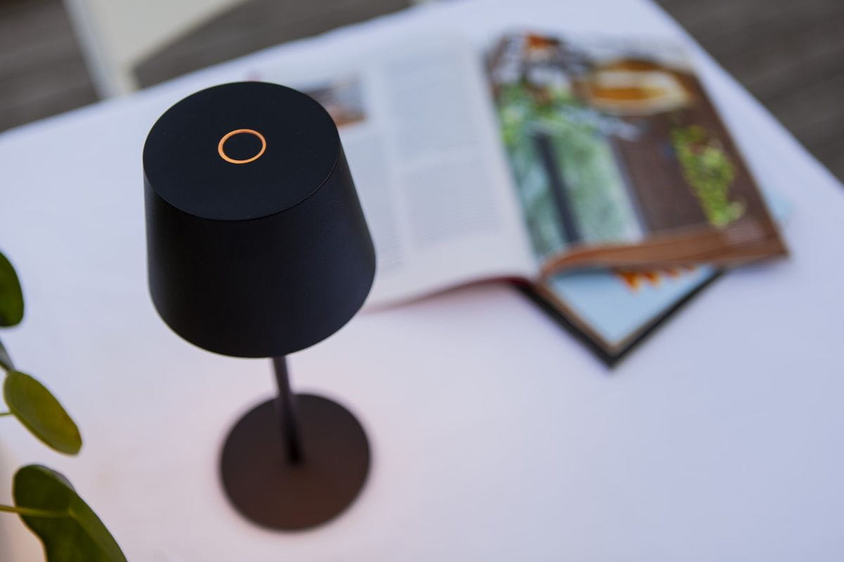 Lampka stołowa ogrodowa Roble w kolorze czarnym, z okrągłym, matowym kloszem i cienką podstawą. Ujęcie z góry, na tle białego stołu i otwartej gazety. Widoczny detal na górze klosza w formie okręgu.