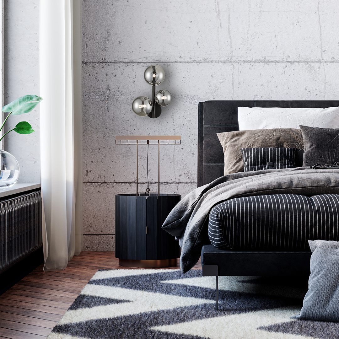 Mila kinkiet 3xG9 w kolorze czarnym, z trzema metalowymi kulami, zamontowany na betonowej ścianie. W tle widoczne nowoczesne łóżko z poduszkami, stolik nocny oraz roślina w doniczce.
