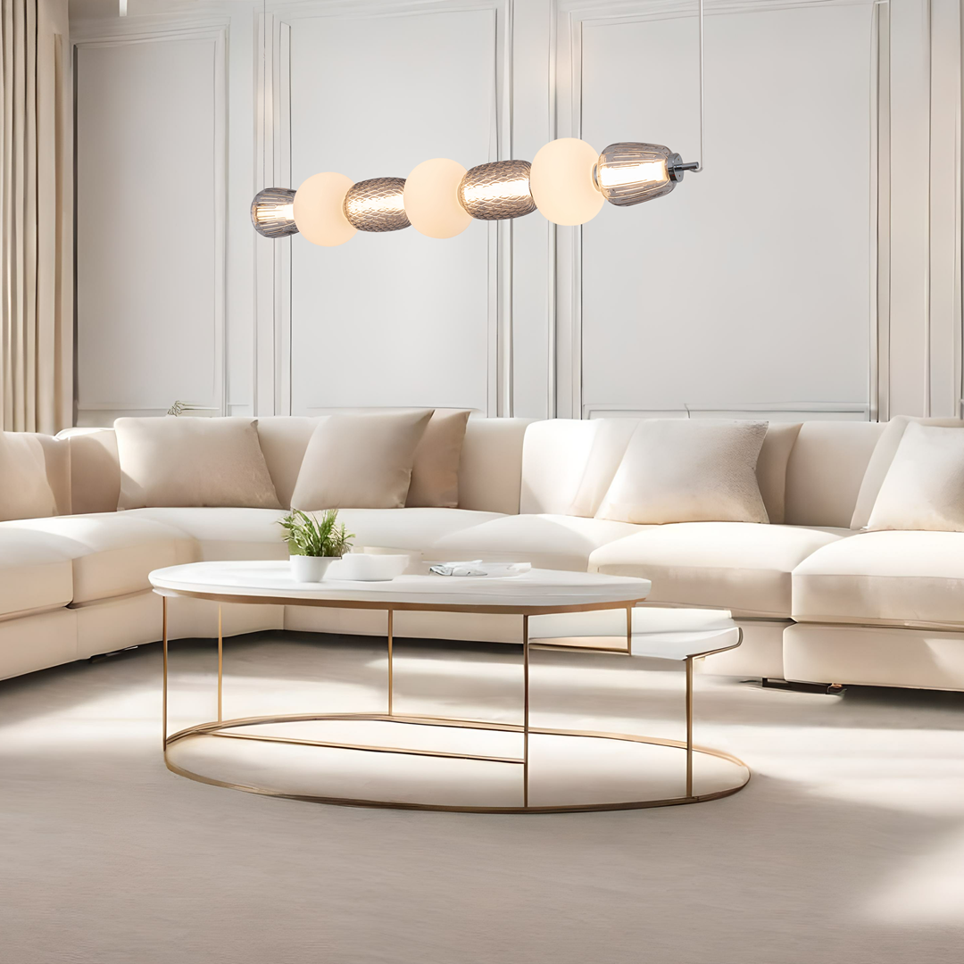 Lampa wisząca Caro w chromowanej obudowie z pięcioma okrągłymi, białymi kloszami, zawieszona nad okrągłym stołem kawowym. Lampa emitująca światło LED 42W, idealna do nowoczesnych wnętrz. Tło w jasnych odcieniach, z elegancką sofą w kolorze ecru.