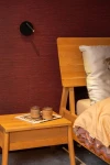 Kinkiet Jolijn w kolorze czarnym, zasilany bateryjnie z jedną diodą LED o mocy 2W. Ujęcie z boku, zamontowany na ścianie przy łóżku, obok drewnianej szafki nocnej z dwoma filiżankami i książką na stole. Tło w odcieniu ciemnego czerwonego.