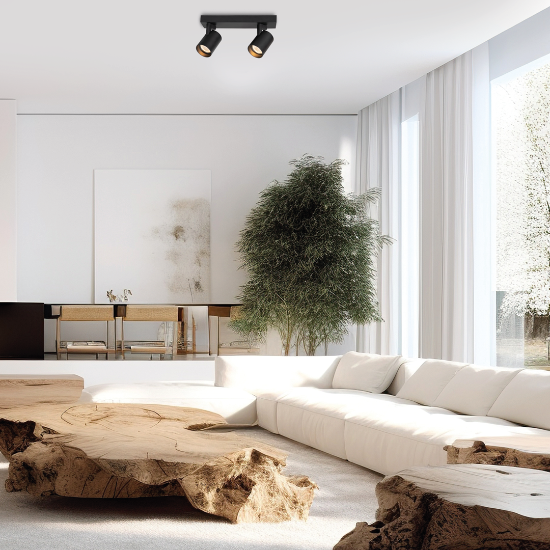 Czarna lampa sufitowa Volf z dwoma reflektorami GU10, zamontowana na suficie, w nowoczesnym wnętrzu. W tle widoczna jasna kanapa oraz drewniany stół. Otoczenie jasne, z dużymi oknami i roślinnością.