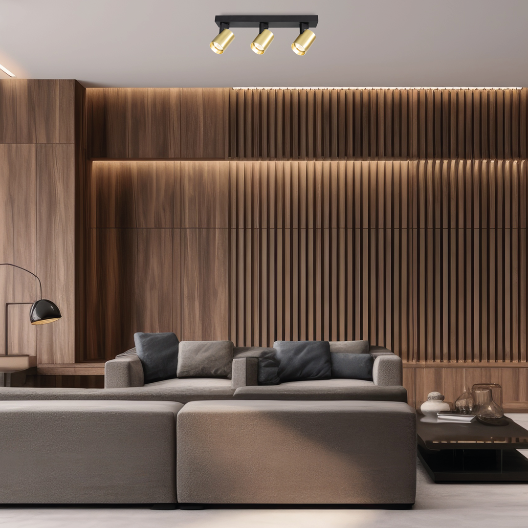 Lampa sufitowa Volf z trzema reflektorami w kolorze czarnym i złotym, zamontowana na prostokątnej podstawie. Reflektory skierowane w dół, w nowoczesnym wnętrzu z drewnianą ścianą i sofą w odcieniach szarości.