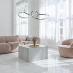 Lampa wisząca Marco LED w kolorze chromu, o nowoczesnym, niesymetrycznym kształcie z dwoma pętlami, zawieszona na cienkich linkach. Otoczenie to jasny salon z miękkim, różowym narożnikiem i okrągłym fotelem, na stole znajduje się złota taca z wazonem.