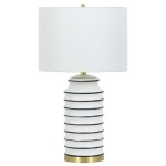 Lampa stołowa Coastal z białym abażurem i podstawą z mosiądzu, w kształcie cylindrycznym z poziomymi, ciemnoniebieskimi paskami. Stoi na płaskiej powierzchni, z widocznym detalem w postaci złotego wykończenia u podstawy.