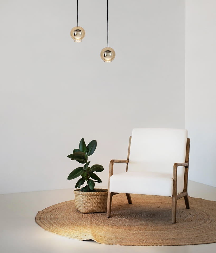Lampa wisząca Ojo z jedną żarówką LED 7W, w złotym kolorze, zawieszona na czarnych przewodach. W tle widoczny biały fotel z drewnianymi ramami oraz roślina w koszu na okrągłym dywaniku. Proste, minimalistyczne tło.