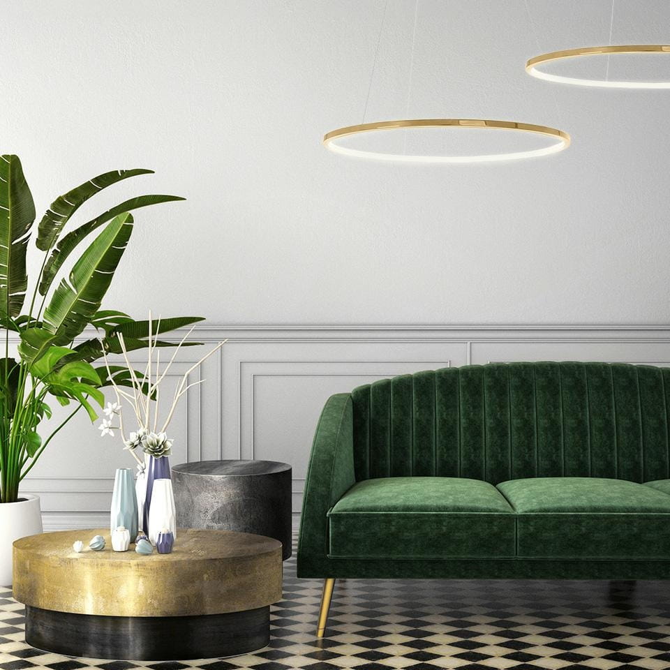 Złota lampa wisząca Ring Slim 80 z pilotem, wyposażona w jedno źródło światła LED o mocy 28W. Produkt zawieszony w przestrzeni, nad okrągłym, złotym stolikiem, obok zielonej sofy i roślinności w nowoczesnym wnętrzu.