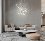 Lampa wisząca Ring Slim S w złotym kolorze, z trzema okrągłymi pierścieniami LED, zawieszona nad okrągłym stołem w nowoczesnym wnętrzu. W tle szara ściana i minimalistyczne meble. Pilot do zdalnego sterowania widoczny obok.
