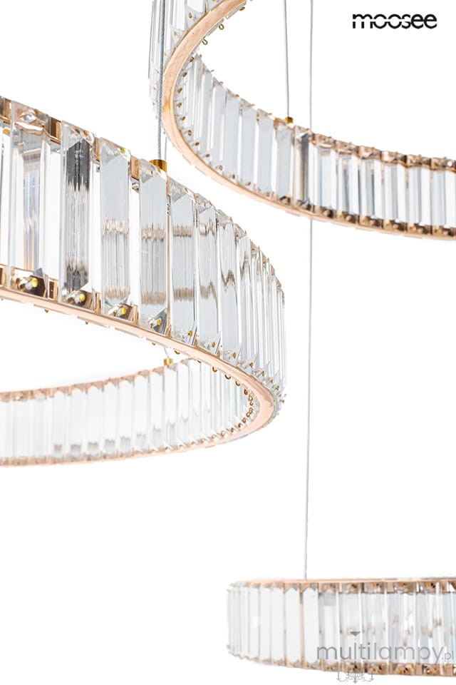 Lampa wisząca Liberty 3B w złotym kolorze, z trzema źródłami światła LED, posiada okrągłe pierścienie zdobione przezroczystymi kryształami. Widoczny detal to metalowe wykończenie i sznurki montażowe. Produkt wisi w powietrzu na białym tle.
