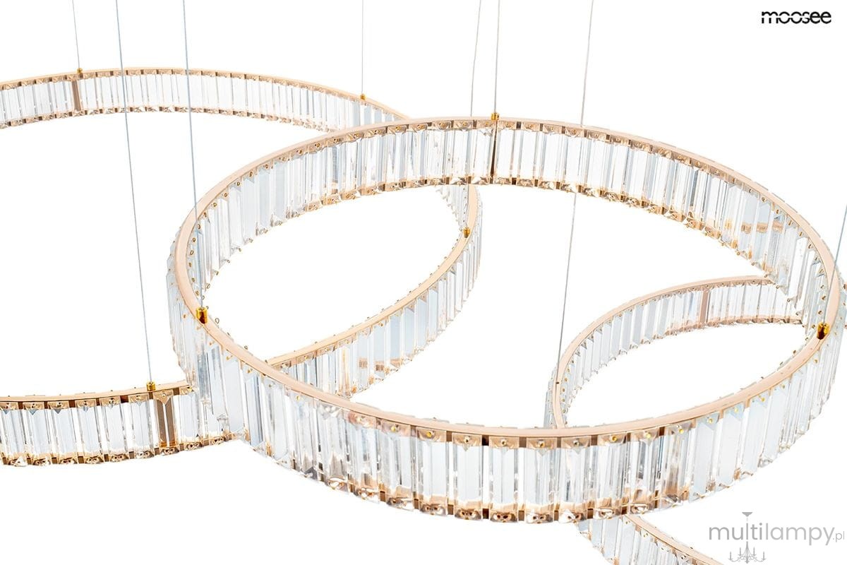 Lampa wisząca Liberty 3B w kolorze złotym, z trzema źródłami LED. Okrągłe pierścienie z transparentnego szkła zdobione kryształkami. Produkt zawieszony na cienkich linkach, pokazany z bliska, podkreślający detal i fakturę. Tło białe.