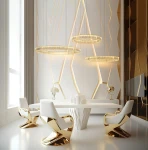 Lampa wisząca Liberty 3B w kolorze złotym, z trzema okrągłymi kloszami LED, zawieszona nad eleganckim stołem w nowoczesnym wnętrzu. Tło z geometrycznymi wzorami w odcieniach bieli i złota, z białymi krzesłami o złotych detalach.