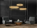Lampa wisząca Liberty 3B z trzema okrągłymi kloszami LED w złotym kolorze, zawieszona w równych odstępach. W tle ciemna ściana oraz nowoczesne, szare fotele obok stołu z drewnianą podstawą i marmurowym blatem.