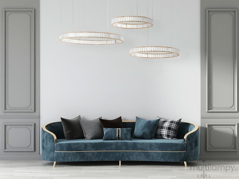 Lampa wisząca Liberty 3B w kolorze złotym, z trzema okrągłymi kloszami LED, zawieszona nad niebieską sofą z poduszkami. Lampa ma nowoczesny design, a tło jest jasne, gładkie, co podkreśla elegancję produktu.
