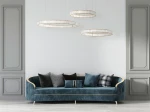 Lampa wisząca Liberty 3B w kolorze złotym, z trzema okrągłymi kloszami LED, zawieszona nad niebieską sofą z poduszkami. Lampa ma nowoczesny design, a tło jest jasne, gładkie, co podkreśla elegancję produktu.