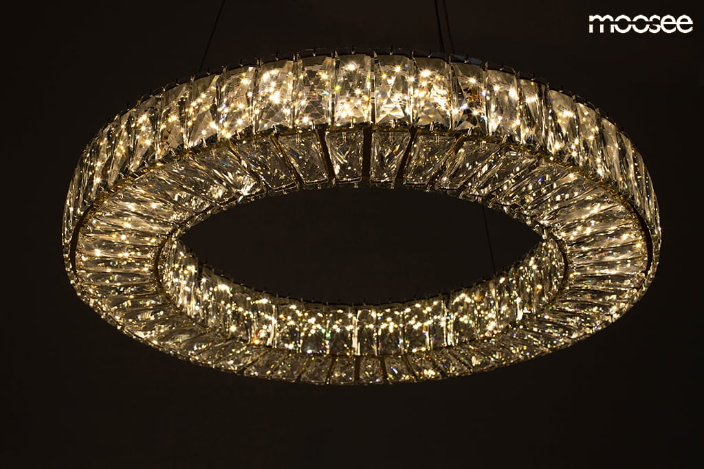 Złota lampa wisząca Allisia 80 z kryształowym kloszem, o okrągłym kształcie. Widoczna jest dioda LED o mocy 43W, która emituje jasne światło, tworząc efektowne odbicia. Lampa zawieszona jest na cienkich linkach w ciemnym otoczeniu.