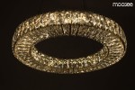 Złota lampa wisząca Allisia 80 z kryształowym kloszem, o okrągłym kształcie. Widoczna jest dioda LED o mocy 43W, która emituje jasne światło, tworząc efektowne odbicia. Lampa zawieszona jest na cienkich linkach w ciemnym otoczeniu.