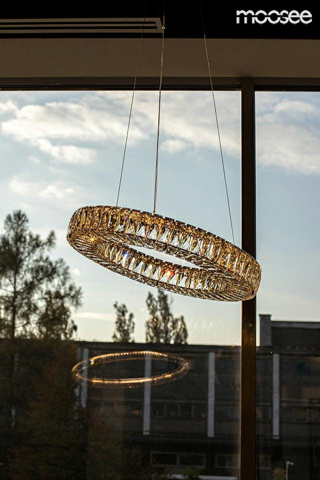 Lampa wisząca Allisia 80 o złotej oprawie, z pojedynczym źródłem światła LED 43W. Okrągła konstrukcja z kryształowymi detalami, zawieszona nad oknem, w otoczeniu drzew i budynków.