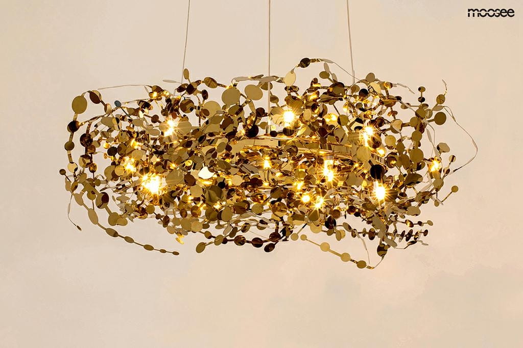 Lampa wisząca Monete 60, złota, o okrągłym kształcie, z 12 źródłami światła G4. Wykonana z metalowych, dekoracyjnych elementów w formie okrągłych, błyszczących detali, zawieszona na cienkich linkach w jasnym otoczeniu.