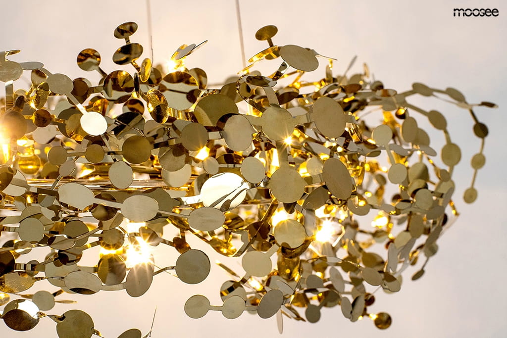 Lampa wisząca Monete 60 z 12 źródłami światła G4, w złotym kolorze. Ozdobna konstrukcja z metalowych, okrągłych elementów w różnych rozmiarach, tworząca efektowną aranżację. Zawieszona w górnej części, na jasnym tle.