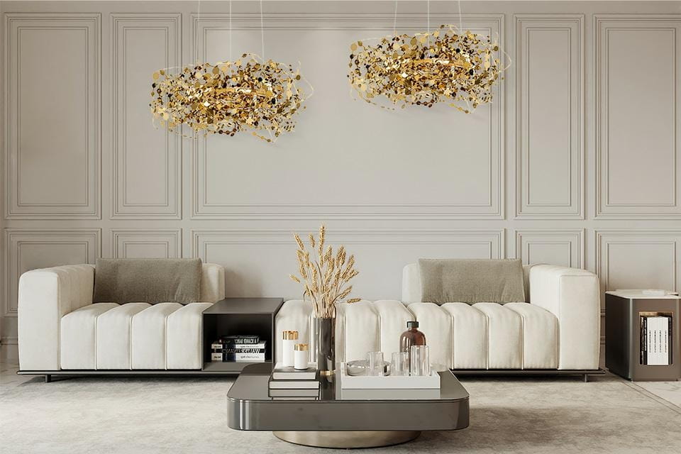 Lampa wisząca Monete 60 w kolorze złotym, z 12 źródłami światła G4, zawieszona w eleganckim wnętrzu. W tle widoczne jasne sofy i stół kawowy. Lampa ma organiczny kształt z dekoracyjnymi elementami przypominającymi liście.