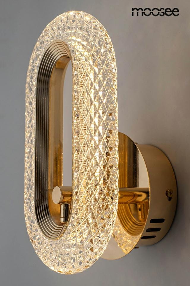 Kinkiet Jewel w kolorze złotym z diamentowym wzorem. Owalna lampa LED o mocy 7W, montowana na ścianie, z metalowym wykończeniem. Świeci ciepłym światłem, tworząc elegancką atmosferę. Tło w neutralnym odcieniu.